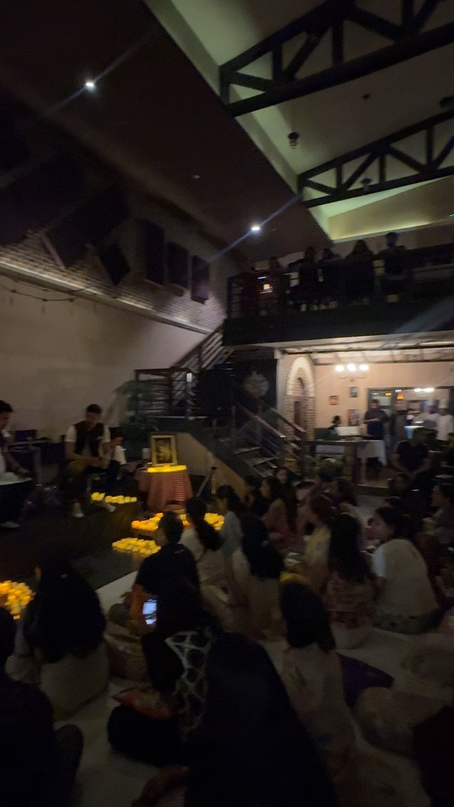 The wider room, the audience seated on the floor facing the marigold-lined stage.